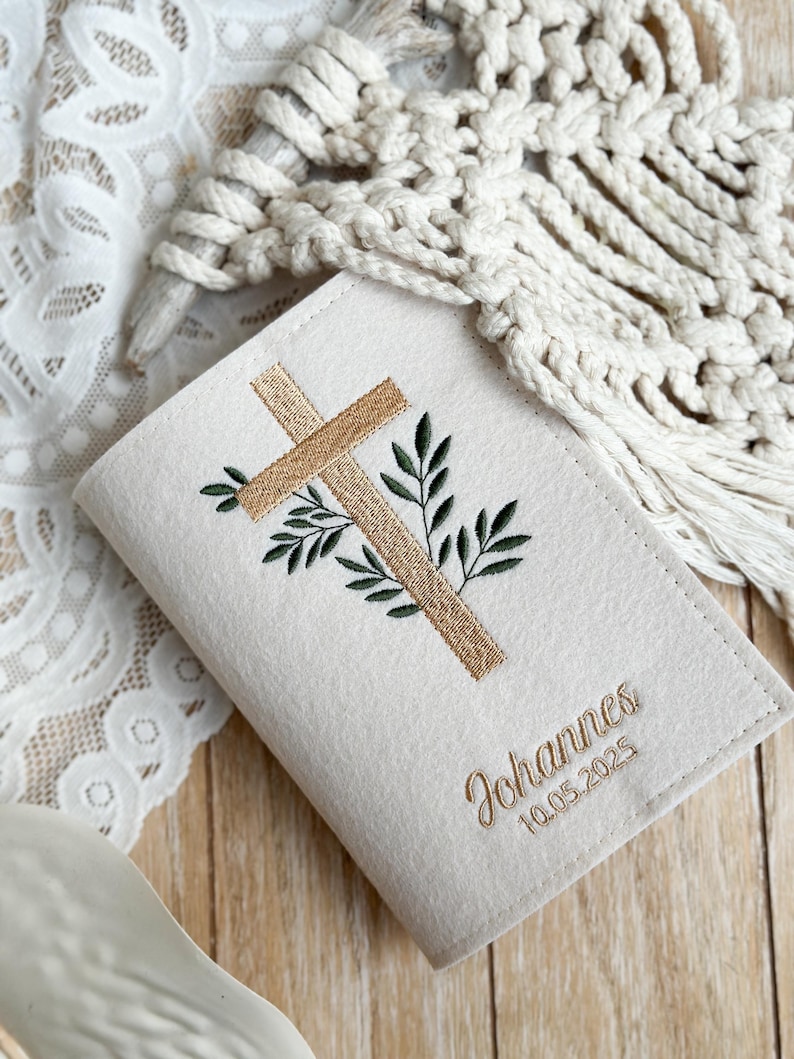 May include: A white felt book cover with an embroidered cross and green leaves. The text "Johannes 10.05.2025" is embroidered below the cross.