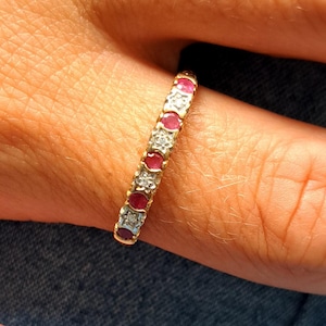 May include: A gold ring featuring alternating red gemstones and clear diamonds. The ring is worn on a finger, highlighting the detailed design and sparkle. The ring is a classic eternity band style.