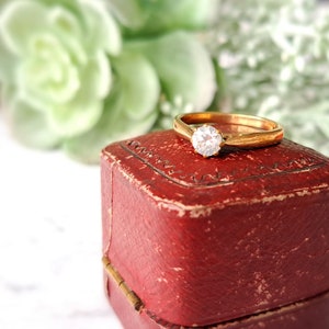 May include: A gold engagement ring with a single round diamond, resting in a red velvet ring box.