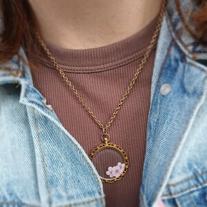 May include: A gold-colored necklace with a round pendant containing small pink flowers. The pendant hangs from a delicate chain. The person is wearing a brown shirt and a denim jacket.