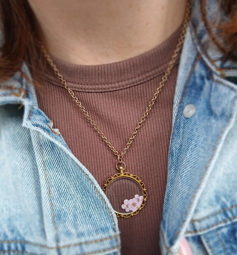 May include: A gold-colored necklace with a round pendant containing small pink flowers. The pendant hangs from a delicate chain. The person is wearing a brown shirt and a denim jacket.