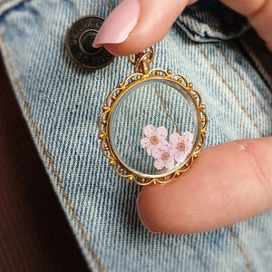 May include: A gold-colored, round pendant necklace with a clear glass center, showcasing delicate pink flowers. The pendant has an ornate, scalloped edge. The necklace is held against a denim jacket.