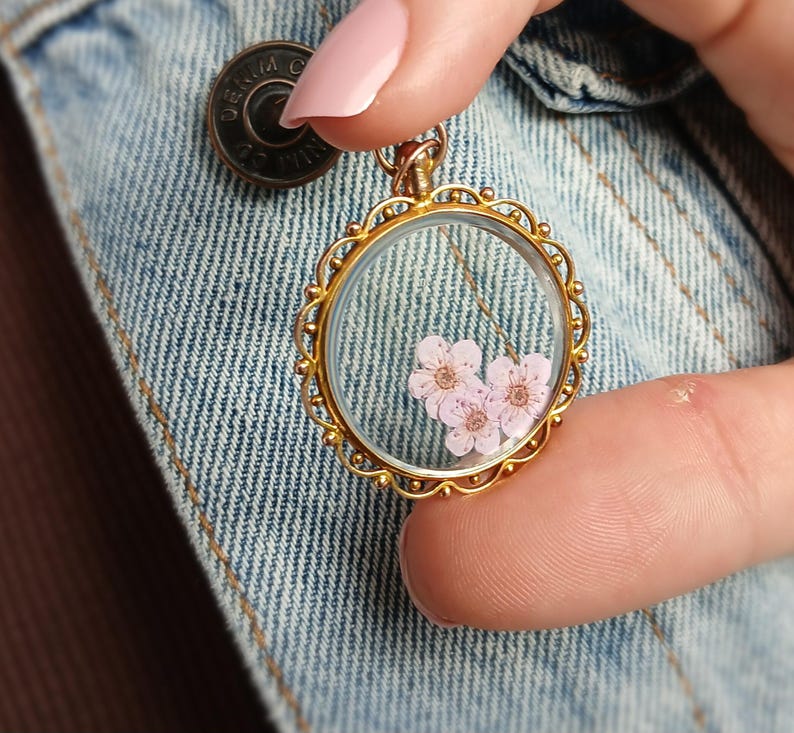May include: A gold-colored, round pendant necklace with a clear glass center, showcasing delicate pink flowers. The pendant has an ornate, scalloped edge. The necklace is held against a denim jacket.
