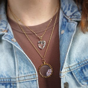 May include: A layered necklace set featuring three gold chains. The top chain holds a clear, heart-shaped pendant. The middle chain has a simple bar pendant. The bottom chain has a round, gold-framed pendant with small pink flowers.