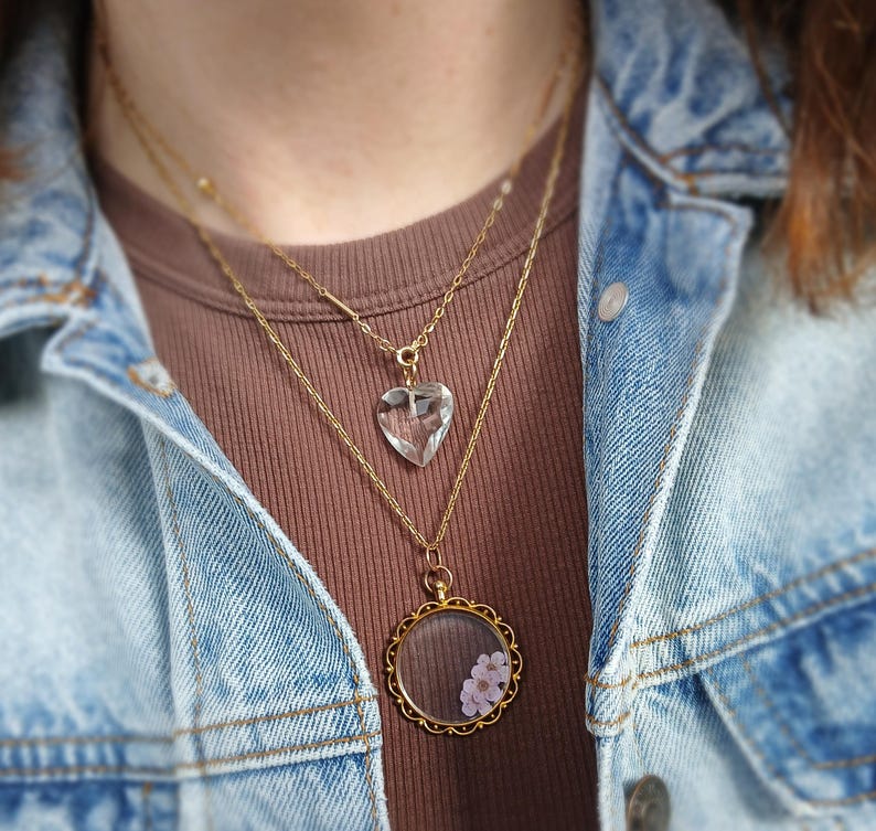 May include: A layered necklace set featuring three gold chains. The top chain holds a clear, heart-shaped pendant. The middle chain has a simple bar pendant. The bottom chain has a round, gold-framed pendant with small pink flowers.