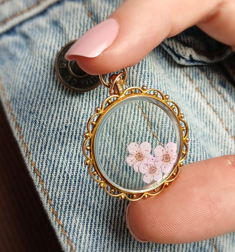 May include: A gold-colored, round pendant necklace with a clear glass front, showcasing delicate pink flowers. The pendant has a decorative, scalloped edge and is suspended from a thin chain. The background is a denim jacket.