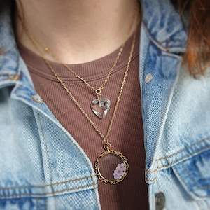 May include: A layered necklace set featuring two gold chains. The top chain holds a clear, heart-shaped pendant. The lower chain has a round, gold-framed pendant with small pink flowers inside. The necklaces are worn over a brown shirt and a blue denim jacket.