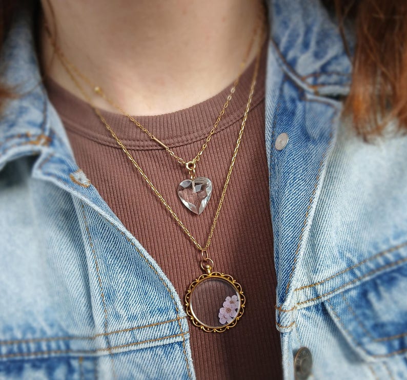 May include: A layered necklace set featuring two gold chains. The top chain holds a clear, heart-shaped pendant. The lower chain has a round, gold-framed pendant with small pink flowers inside. The necklaces are worn over a brown shirt and a blue denim jacket.
