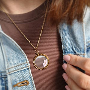 May include: A gold-colored necklace with a round pendant. The pendant has a clear glass center with a small pink flower inside. The necklace is worn over a brown shirt and a blue denim jacket.