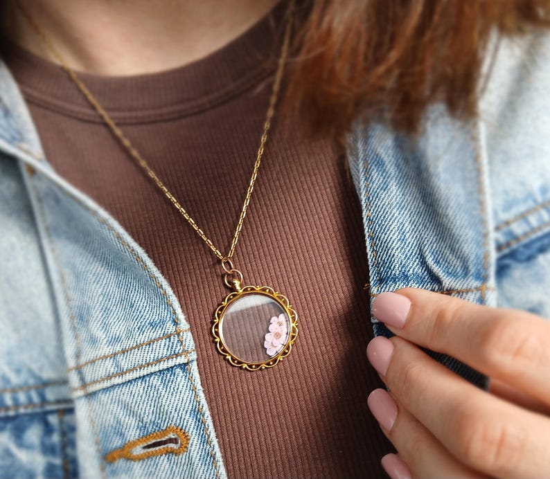May include: A gold-colored necklace with a round pendant. The pendant has a clear glass center with a small pink flower inside. The necklace is worn over a brown shirt and a blue denim jacket.