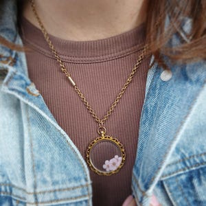 May include: A gold-colored necklace with a round pendant containing small pink flowers. The pendant has a decorative, scalloped edge. The necklace is worn over a brown ribbed shirt and a blue denim jacket.