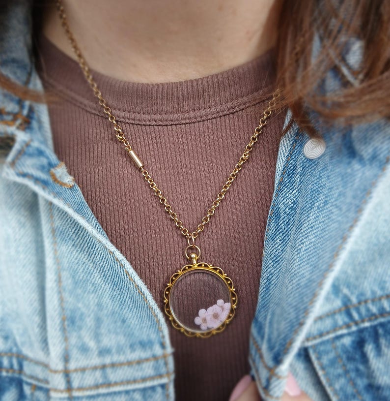 May include: A gold-colored necklace with a round pendant containing small pink flowers. The pendant has a decorative, scalloped edge. The necklace is worn over a brown ribbed shirt and a blue denim jacket.