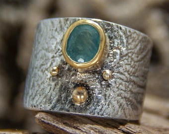 May include: A silver ring with a textured finish and a green gemstone set in a gold bezel. The ring has three small gold accents.