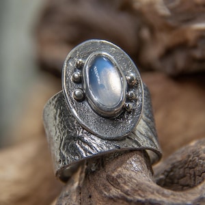 以下が含まれることがあります： テクスチャ加工されたバンドと、白いムーンストーンがセットされた丸いベゼルが付いたシルバーリング。ベゼルは小さなシルバービーズで飾られています。