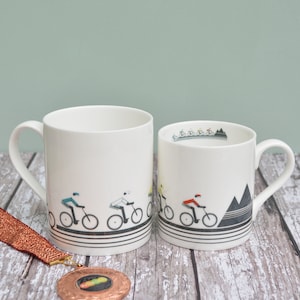 May include: Two white ceramic mugs with black and white cycling themed designs. The larger mug features a design of cyclists riding bikes on a road. The smaller mug features a design of cyclists riding bikes on a road with mountains in the background.