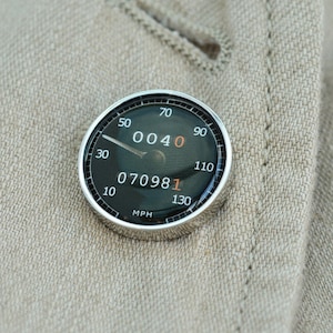 May include: A silver-colored circular pin with a black face and white numbers. The pin has a needle pointing to the number 40. The face also displays the numbers 070981. The pin reads "MPH" at the bottom.