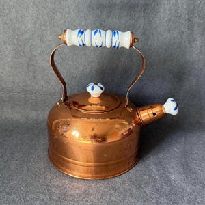 Puede incluir: Una tetera de cobre con un asa y un pico con diseños blancos y azules. La tapa también tiene un pomo blanco y azul. La tetera es redonda y tiene un acabado brillante. El asa es de cobre.