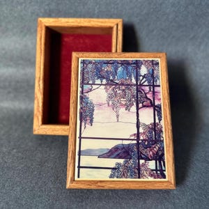 Puede incluir: Una caja de madera con una tapa con bisagras. La tapa presenta una impresión colorida de una glicinia con flores moradas y una escena de paisaje. La caja está cerrada.