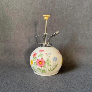 May include: A spherical white ceramic plant mister featuring a colorful floral pattern with pink, yellow, and blue flowers and green foliage. The top assembly is made of polished chrome metal with a small cream-colored plastic plunger handle.