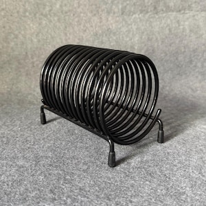 May include: Black metal wire letter rack. The rack features a circular design made of curved wires, supported by a rectangular base with four short legs. The design is minimalist and modern.
