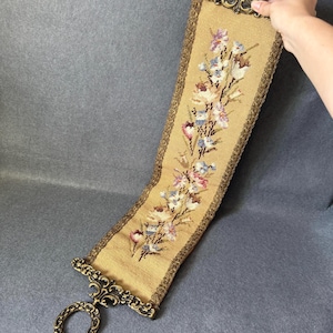 May include: A vintage tapestry wall hanging with a floral design. The tapestry has a tan background with embroidered flowers in pink, blue, and white. It features a decorative gold-coloured metal frame and a loop for hanging.