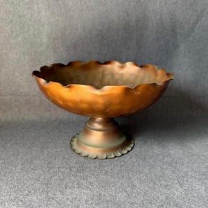 Puede incluir: Un cuenco con pedestal de color cobre con un borde festoneado. El cuenco tiene una base redonda con un borde decorativo festoneado. El interior del cuenco tiene una pátina más oscura, mientras que el exterior es de color cobre brillante.