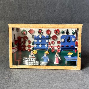 Può includere: Un diorama in miniatura all'interno di una cornice rettangolare in legno che raffigura una scena di cucina. Le pareti sono decorate con numerosi piccoli piatti e padelle colorati. Tre piccole figure si trovano nella stanza con uno scaffale blu.
