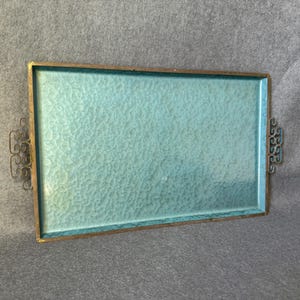 Puede incluir: Bandeja rectangular con superficie azul claro texturizada y marco de metal dorado. La bandeja tiene asas decorativas en cada extremo. Una pieza decorativa de estilo vintage.
