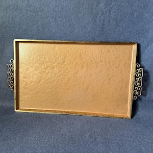 May include: A rectangular serving tray with a textured, copper-colored base and a metallic frame. The tray features ornate, scroll-patterned metal handles on both sides, providing a decorative contrast to the smooth, flat surface.
