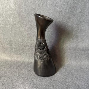 May include: A dark grey vase with an unusual, asymmetrical form. The vase features a flared top and an intricate floral design carved into the lower section. The vase is made of a dark, possibly metallic material.
