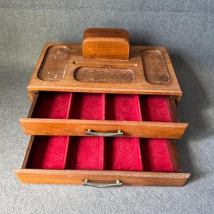 Puede incluir: Caja de joyería de madera con dos cajones extraíbles forrados con terciopelo rojo. La sección superior tiene compartimentos y un pequeño bloque de madera. La caja está hecha de madera de color marrón claro.