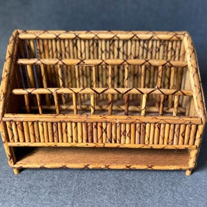 Op de afbeelding: Een kleine, rechthoekige, houten tijdschriftenrek gemaakt van geweven bamboestokken. Het rek heeft een plank aan de onderkant en een decoratief, geweven patroon aan de voorkant.