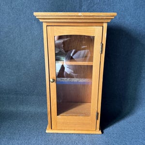 May include: A small, wooden display cabinet with a single glass door. The cabinet is made of light-colored wood and features a small brass knob and a shelf inside. The door is hinged on the right side.