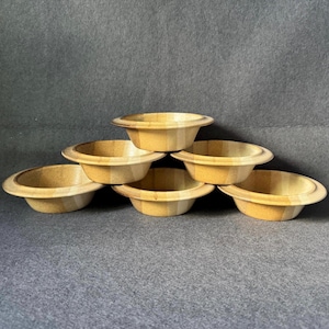Könnte beinhalten: Sechs kleine, runde Holzschalen in hellem Braun sind pyramidenförmig angeordnet. Jede Schale hat einen breiten, flachen Rand und eine etwas tiefere Mitte. Die Schalen scheinen handgefertigt zu sein und haben eine natürliche, rustikale Ästhetik. Jede Schale misst etwa 15 cm im Durchmesser.