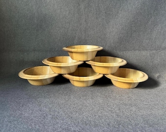 Puede incluir: Un conjunto de seis cuencos pequeños y redondos de madera de color marrón claro dispuestos en forma de pirámide. Cada cuenco presenta un acabado liso y pulido con vetas de madera natural visibles y un borde ancho y plano.