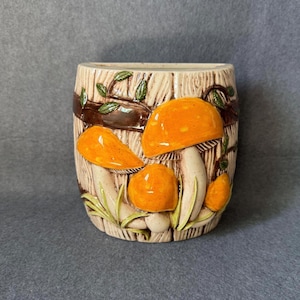 Op de afbeelding: Een keramische wandplantenbak met een reliëfontwerp van feloranje paddenstoelen en groene bladeren tegen een gestructureerde beige achtergrond met houtnerf. De pot heeft een platte achterkant voor montage en een gebogen voorkant.