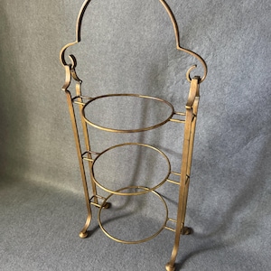 Puede incluir: Un soporte de servicio dorado de tres niveles con un asa arqueada decorativa. El soporte presenta tres plataformas circulares y detalles ornamentados. El soporte de metal está diseñado para exhibir postres u otros artículos.