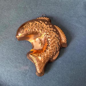 May include: A copper-colored fish-shaped mold with a textured surface. The mold has a loop at the top for hanging and is likely used for making decorative food items. The fish design is detailed and three-dimensional.