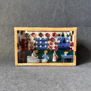 Puede incluir: Un diorama en miniatura que representa una escena de cocina con pequeñas figuras. La escena incluye estantes con platos rojos y negros, armarios azules y mesas pequeñas. Las figuras visten ropa colorida, y una pequeña mesa sostiene un cuenco de fruta.