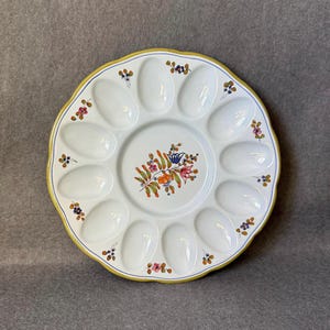 May include: White ceramic egg serving platter with 12 oval indentations and a central floral design. The rim is scalloped with a yellow border and blue trim. Small floral accents are placed around the rim.