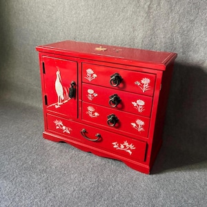 May include: A red lacquered wooden jewelry chest featuring a single door with a white crane inlay and four drawers adorned with white floral inlays. The chest includes dark metal ring pulls on each drawer and a small metal latch on the door.