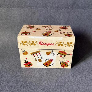 May include: A rectangular cream-coloured metal recipe box featuring the word Recipes in red script. The box is decorated with colourful illustrations of kitchenware including coffee grinders, kettles, spatulas, and fruit in red, black, and gold.