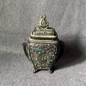 May include: A dark, weathered metal vessel with a lid, featuring intricate relief carvings of swirling patterns and mythical creatures. The container has two side handles, four small feet, and a decorative finial on the lid.