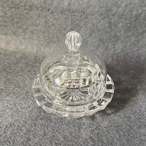 Puede incluir: Una mantequillera de cristal transparente con tapa abovedada y un pomo esférico. La base y la tapa presentan un patrón geométrico de estrella prensado, creando una textura facetada que refleja la luz por toda la superficie del objeto.
