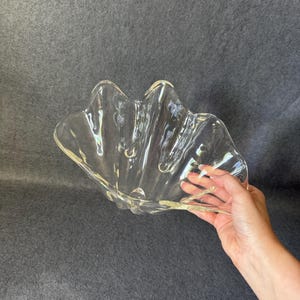 May include: A clear glass bowl with a scalloped, wavy rim and a rippled, shell-like texture. The bowl features a translucent, reflective surface with deep, curved indentations that create a fluid, organic shape throughout the entire piece.