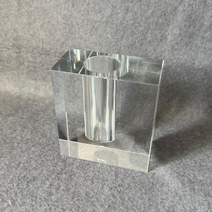 May include: A clear, rectangular acrylic vase with a cylindrical center. The vase has a modern, minimalist design, perfect for displaying flowers or as a decorative accent. The transparent material allows light to pass through, creating a clean, elegant look.