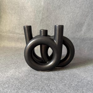 May include: A black ceramic candle holder with a distinctive, interwoven design. It has three vertical cylinders and two circular elements, creating an abstract, modern aesthetic. The matte finish and dark colour give it a minimalist look.