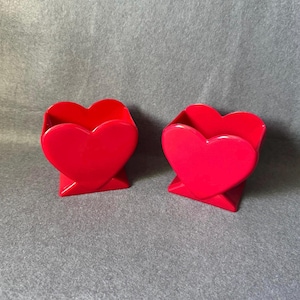 Peut inclure: Deux contenants rouge vif en forme de cœur avec des surfaces lisses et brillantes. Chaque contenant présente une ouverture distincte en forme de cœur sur le dessus et une base stable et inclinée, offrant un aspect uniforme aux deux pièces.