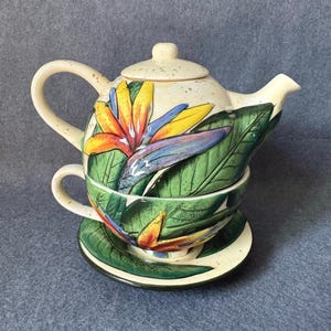 May include: A ceramic teapot, teacup, and saucer set. The cream-colored teapot and cup are decorated with a colorful bird of paradise flower and green leaves. The saucer has a matching design. The set is designed for one person.