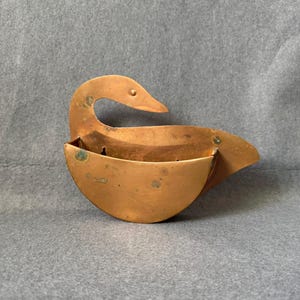 May include: A copper-colored, swan-shaped wall pocket. The swan has a curved neck and a pocket-like body. The metal has a slightly aged appearance with some green patina. The design is simple and elegant, suitable for holding small items.